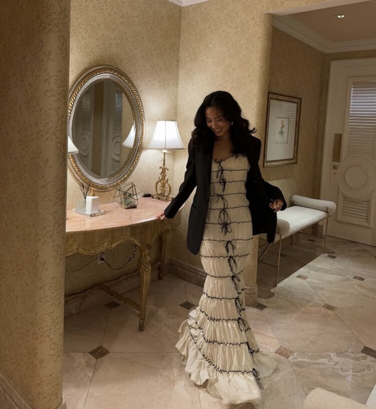 A woman in a long, ruffled beige dress with dark ribbon accents stands in a well-lit, elegant room—perfect for fall date night outfits. She wears a black blazer and smiles while looking down. The room features a circular mirror, ornate lamp, and decorative table.