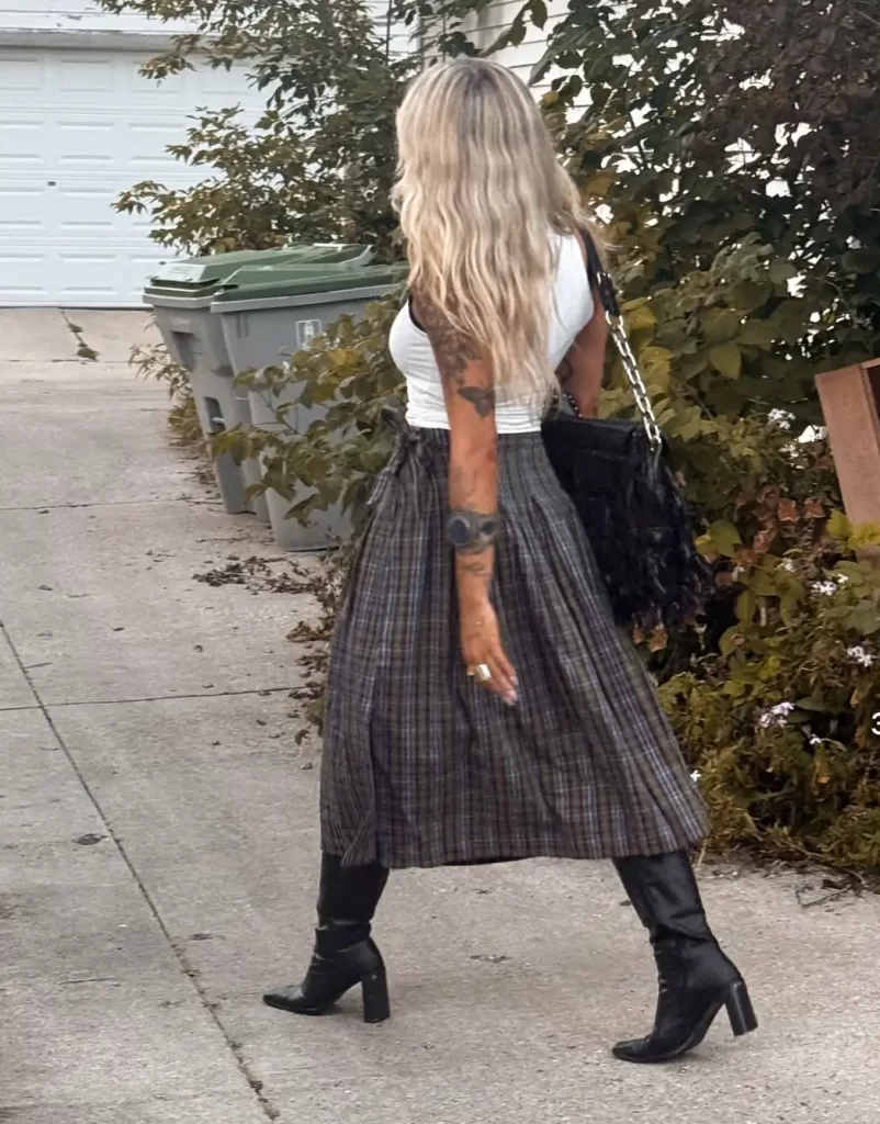 woman walking away wearing long grey plaid skirt with black leather boots and a white tank top