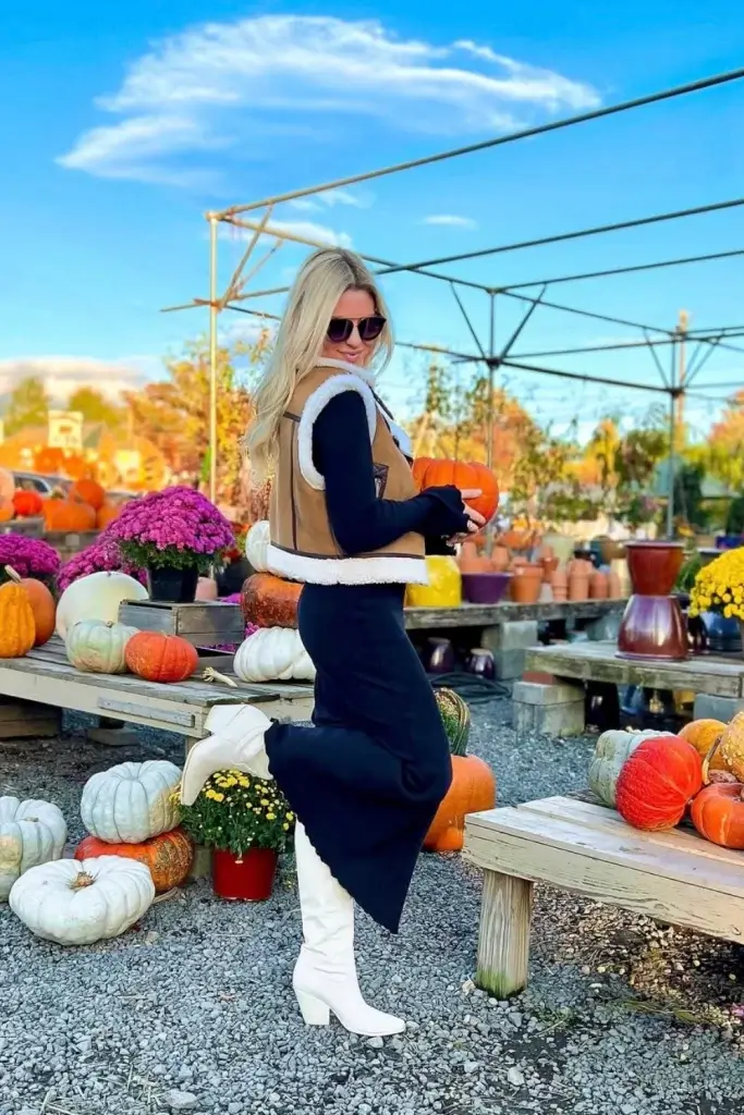 Young blonde woman standing outside near tables of pumpkins wearing a black sweater dress, white boots and a cropped tan sherling vest
