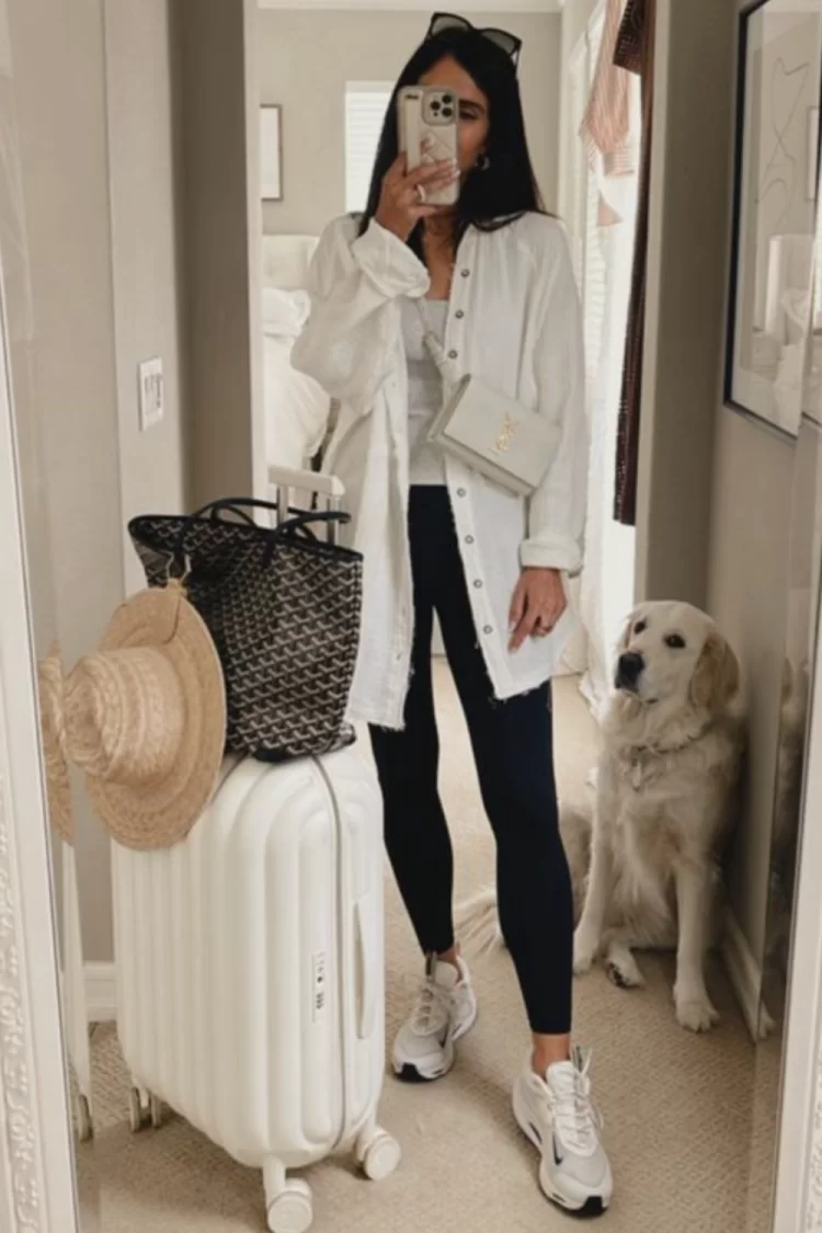 young woman standing in home posing with white luggage and bags getting ready for a trip while she's black leggings, chunky sneakers and a white linen button up