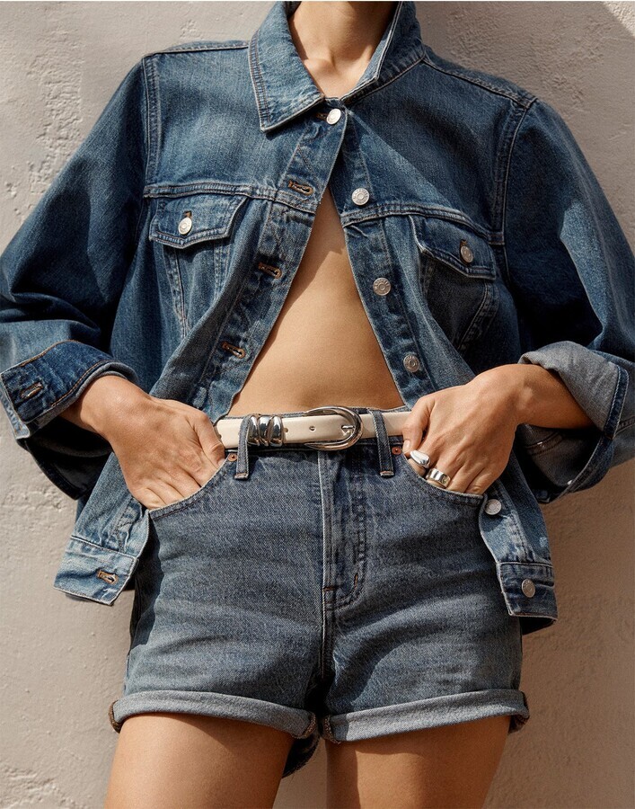 close up on model wearing denim button up top that has top button close showing denim shorts with white leather belt