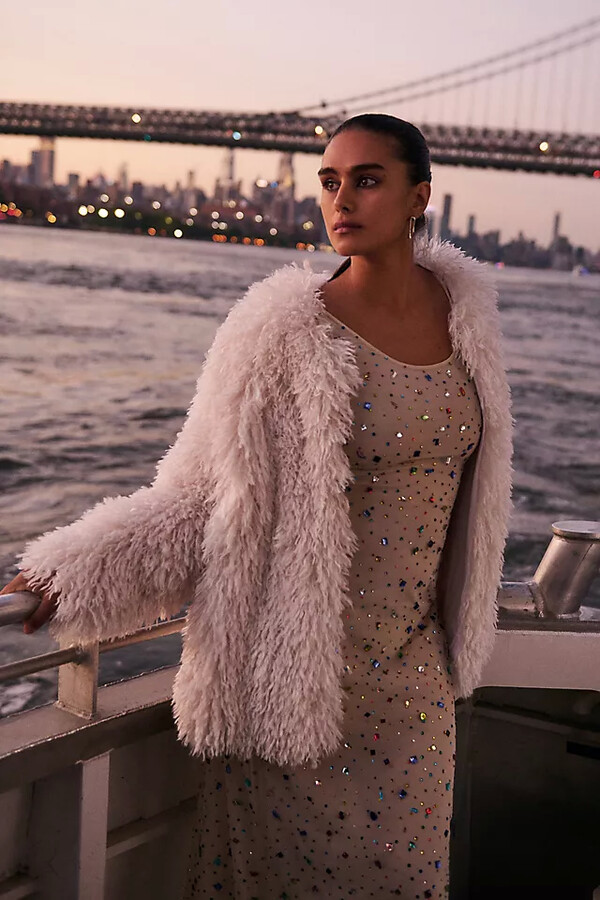 young woman at dusk on a boat overlooking a city landscape and bridge wearing an embellished mesh dress and a shaggy white coat over