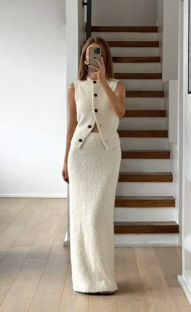 A woman stands indoors, capturing a mirror selfie. She's wearing a cream-colored knit sleeveless top paired with a matching long skirt—an ideal choice for fall bridal shower outfits. The background features a staircase and a minimalistic, modern interior design.