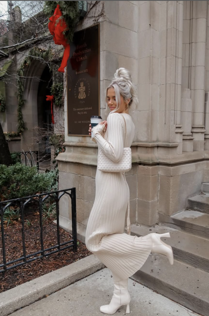 In a white sweater dress and heeled boots, perfect for fall bridal shower outfits, a person stands on a downtown Chicago sidewalk. They hold a cup and smile amidst the winter decorations, with the historic building providing an elegant backdrop.