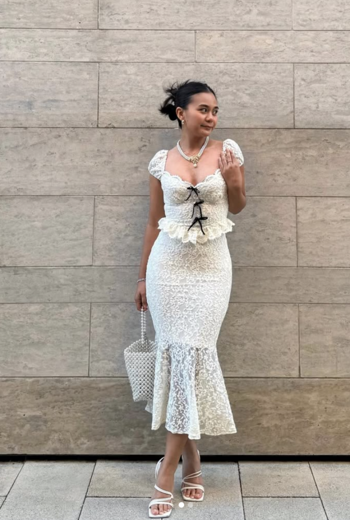 Woman in a white lace dress with a frilly hem, perfect for fall bridal shower outfits, stands against a beige brick wall. She holds a small black purse and wears white heels and a pearl necklace. Her hair is styled up, and she gazes off to the side.