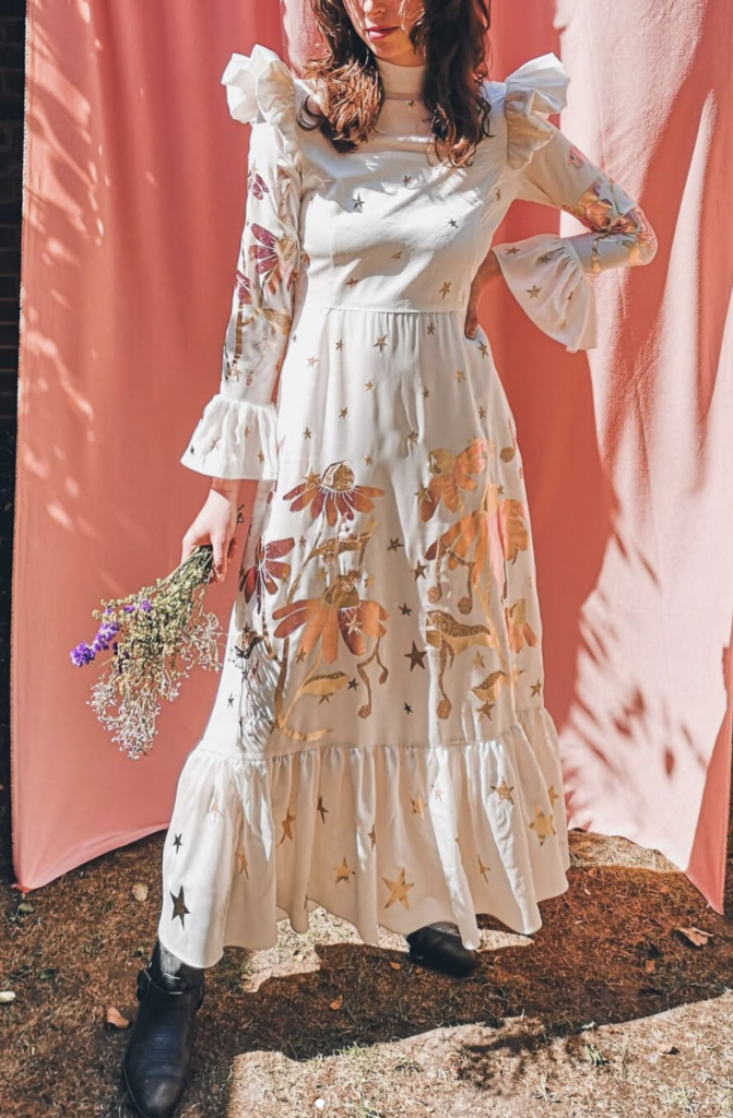 young woman with dark brown hair a white maxi long sleeve dress with ruffles at the shoulder and large metallic motifs throughout standing in front of a pink curtain