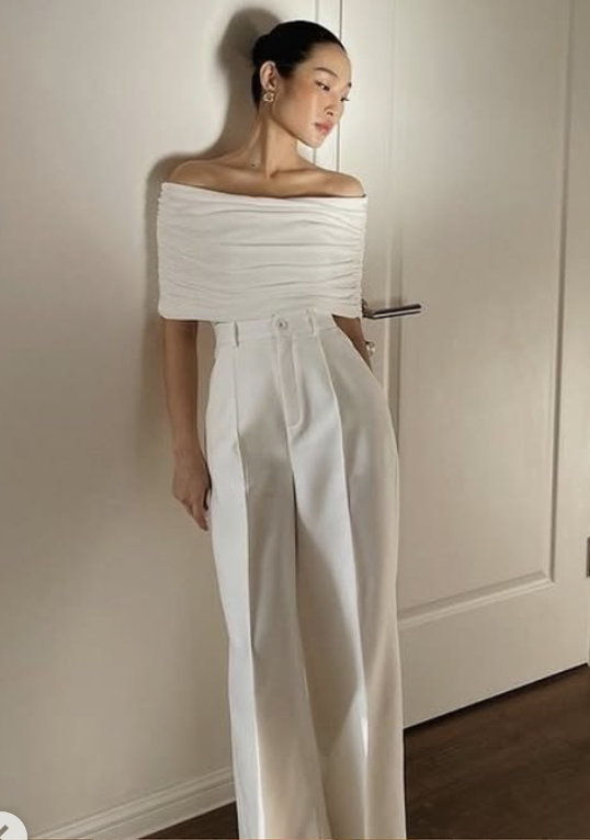 young asian woman leaning against a white wall wearing a mesh off the shoulder top and white trousers