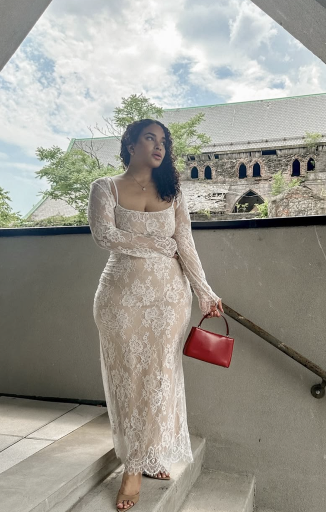 young latin woman posed on a stairway outside wearing a long sleeve lace maxi dress looking off to the side carrying a small red bag