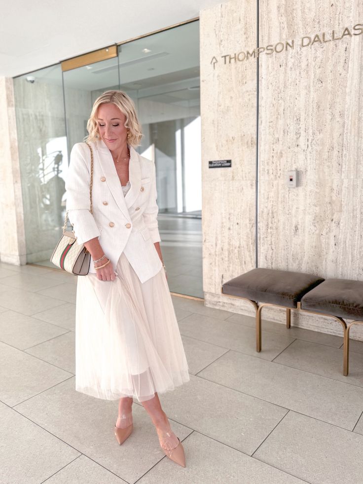 blonde woman posing in front of glass door wearing white blazer, white tulle skirt and nude heels