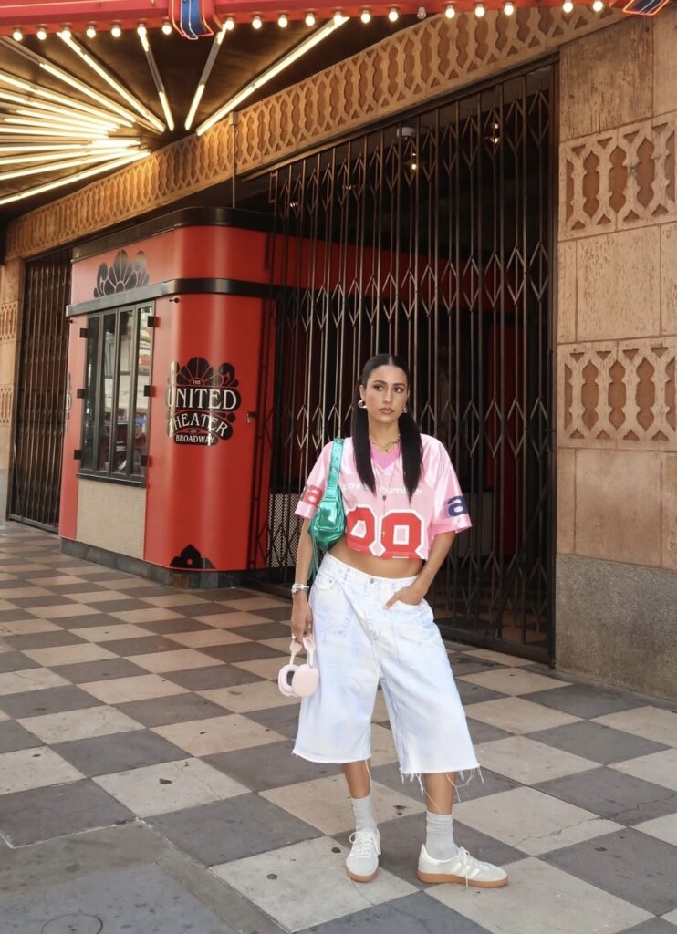 young woman standing in front of theater wearing a cropped pink jersey and oversized baggy white shorts and white sneakers