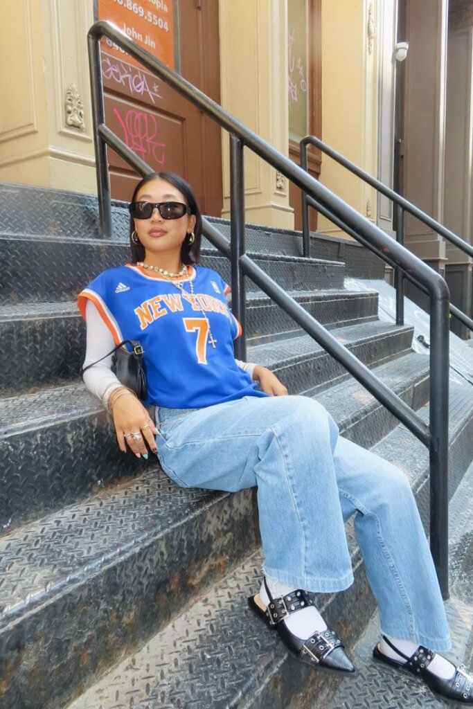 woman leaning against concrete stairs wearing jeans and basketball jersey and black flats