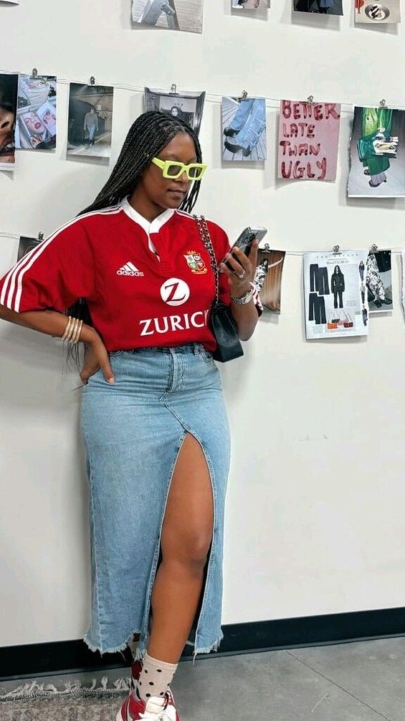 young african american woman standing against wall of photos wear red sports jersey tucked into a denim midi skirt