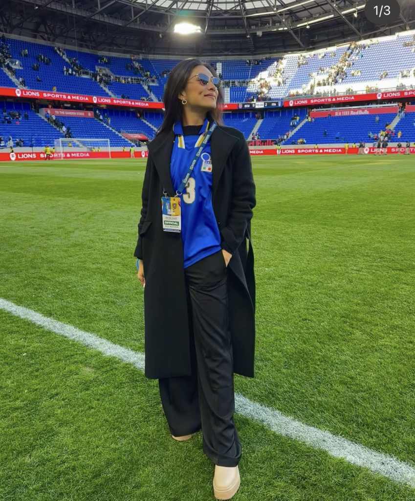 woman standing in football standium wearing blue jersey and black wool coat with black pants and uggs