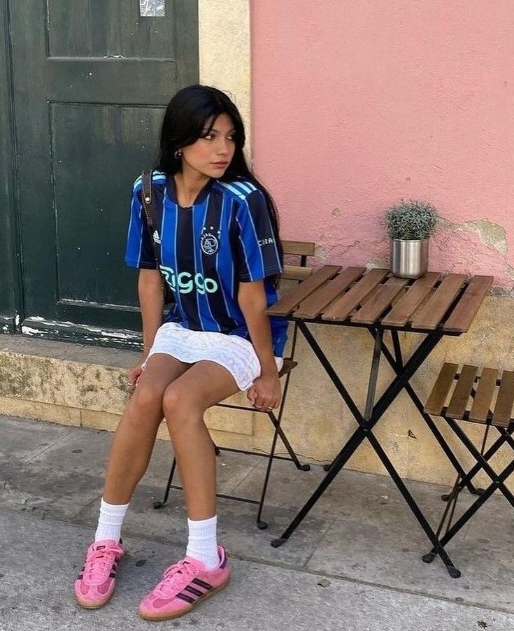 young woman sitting on chair outside wearing pink adidas, white crew socks, white skirt and blue and black striped jersey