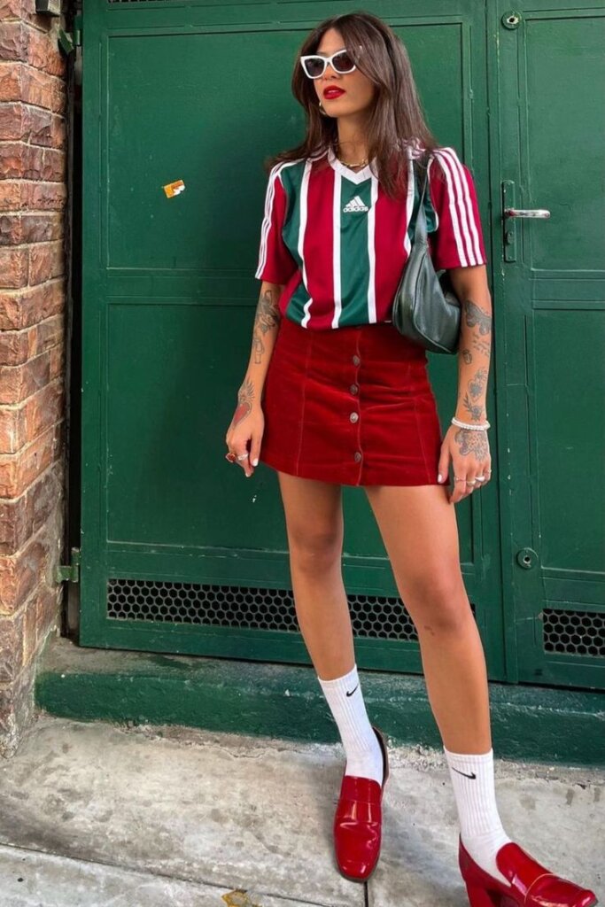 woman standing against green door wear red and green soccor jersey and matching red mini skirt with red shoes.