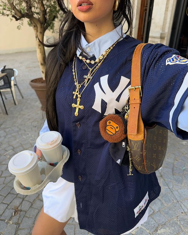 young woman wearing NY sports jersey over a blue button up shirt layered with many gold necklaces holding coffee