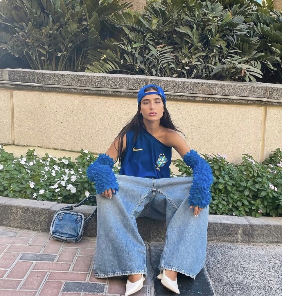 young woman sitting wearing baggy jeans, asymmetrical jersey and matching blue arm warmers and backwards hat