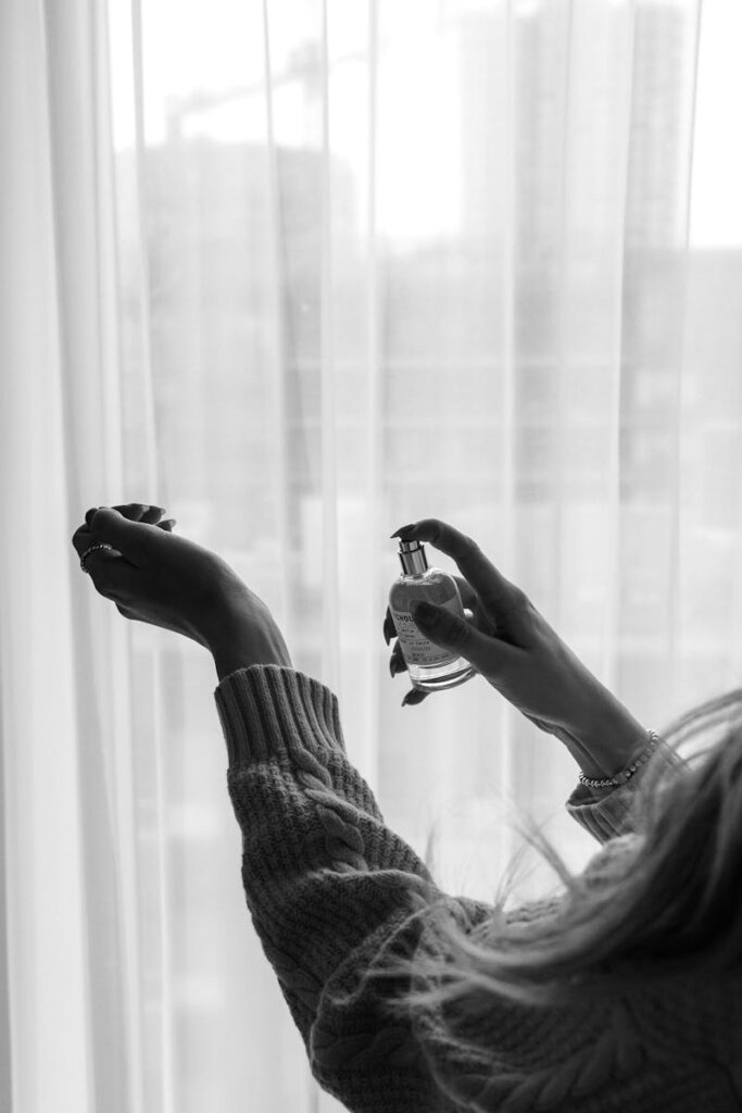 A black and white photo of a woman spraying perfume indoors near a window.