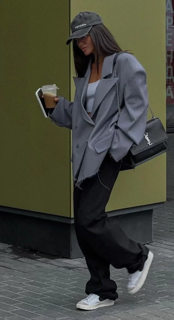 young woman walking down street wearing black sweatpants with white sneakers, grey blazer and green baseball cap