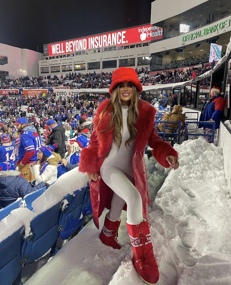 young woman wearing white full length bodysuit and red faux fur coat and moon boots to winter football game