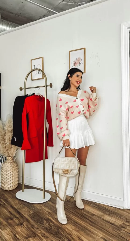 young woman posing in front of rack of clothes wearing a pink hear sweater and white skirt with white boots Valentines day outfit winter