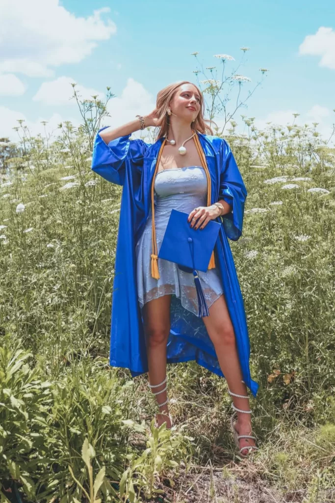 young graduate woman wearing blue graduation gown open to reveal light blue mini dress underneath standing in grassy field