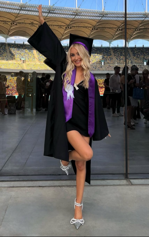 young blonde graduate wearing short black dress and sparkly heels wearing black cap and robe and purple sash