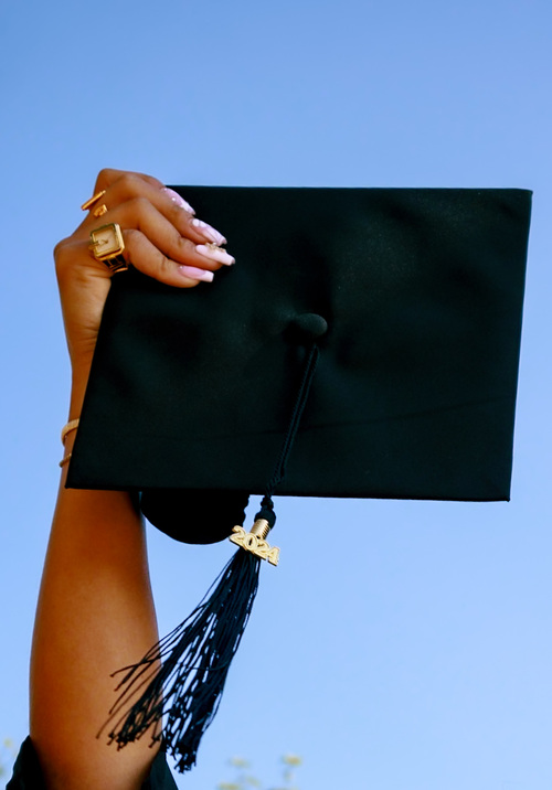 Woman's hand holding up black graduation cap and tassel against sky