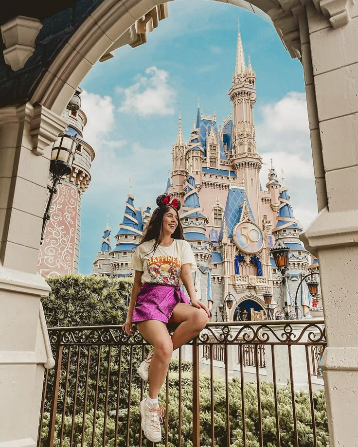 young woman posing on iron fence with Magic Kingdom castle in background as she's wearing metallic purple mini skirt, graphic tee and mickey mouse ears
