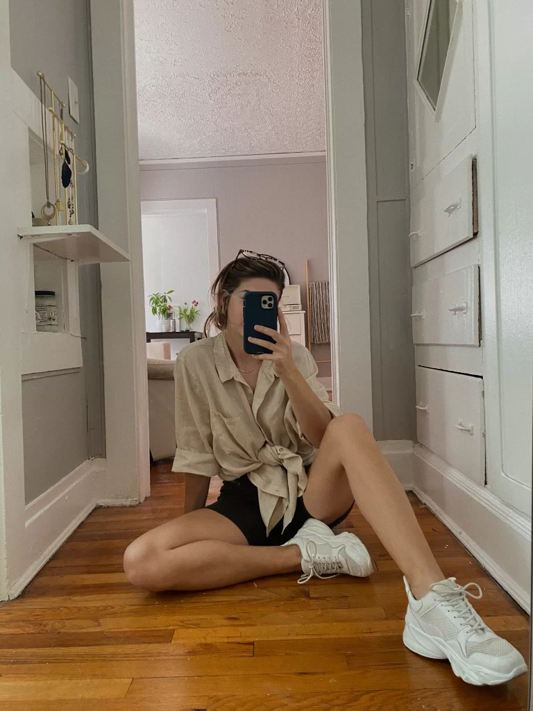 woman sitting on wood floor taking selfie covering face as she shows off her casual summer outfit with chunky white sneakers