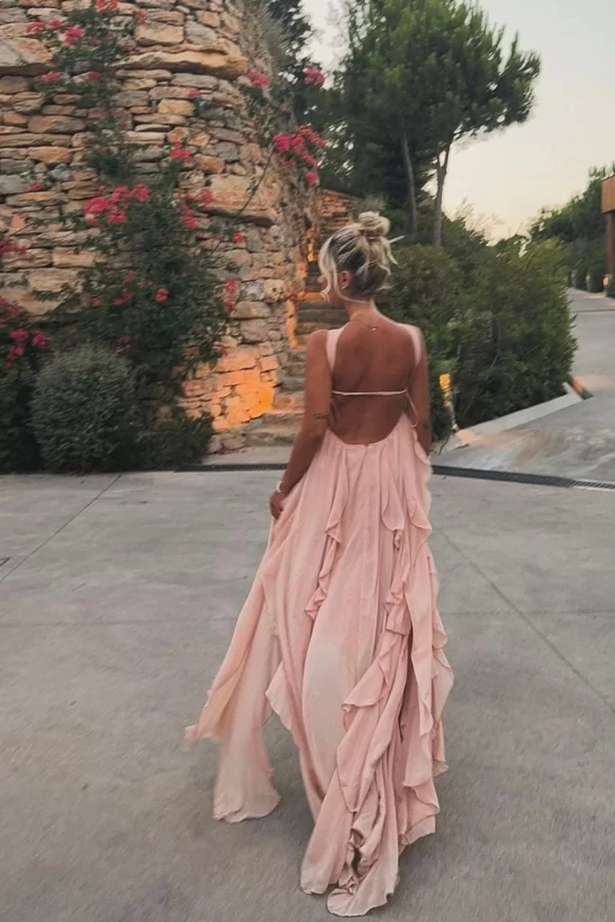 Young blonde woman wearing a peach tone flowy and ruffly boho style backless dress walking off so we see her back