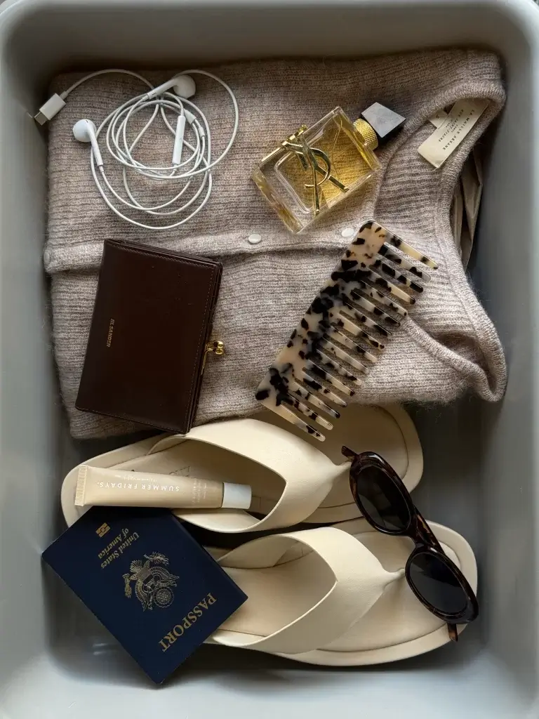 overhead shot of essential travel items packed including light sweater, airpods, fragrance, sunglasses, sandals and passport