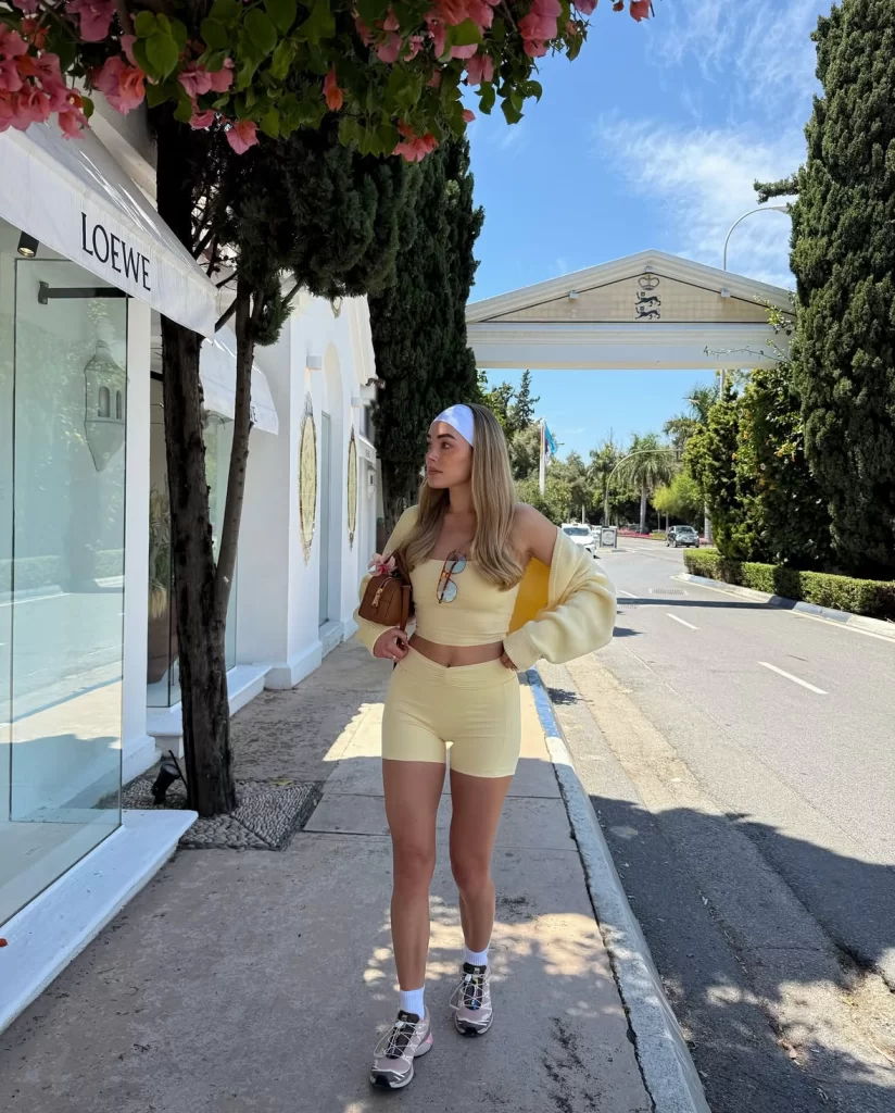 young blonde woman walking down street next to designer stores wearing yellow athleisure set and light pink sneakers