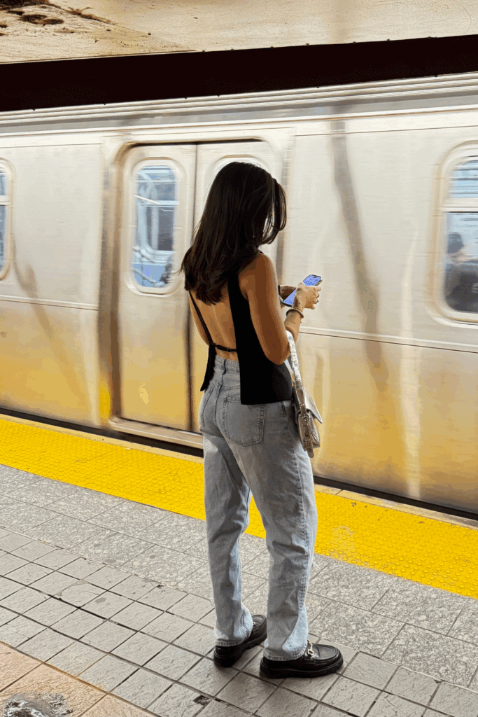 young woman standing in subway terminal wearing black open back top, jeans and black loafers