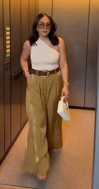 woman walking out of elevator wearing chic and classy outfit for summer of olive color wide leg pants, fitted one shoulder top and a statement belt