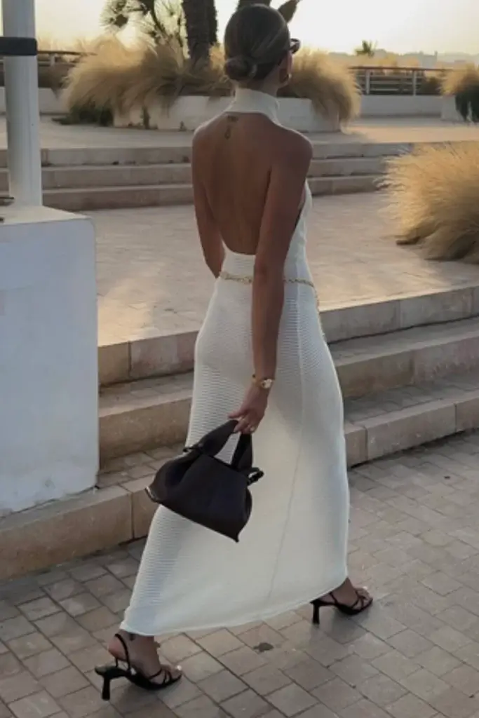 woman walking arcoss decking with beach background wearing an all white open back knit maxi dress with black bag and heels