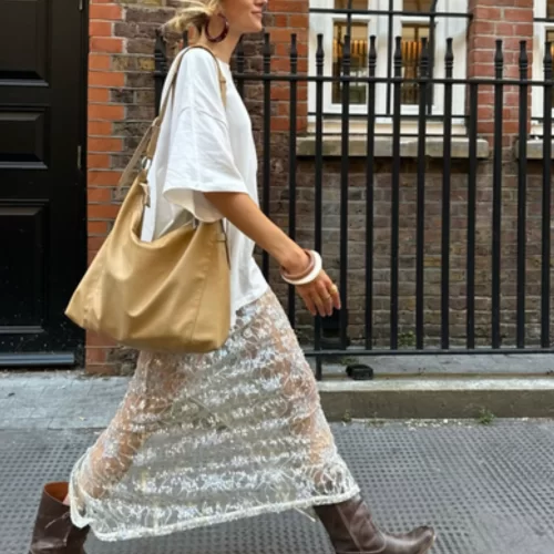 blonde woman walking down street wearing brown leather boots, sequin sheer maxi skirt with an oversized white tee and slouchy leather hobo bag
