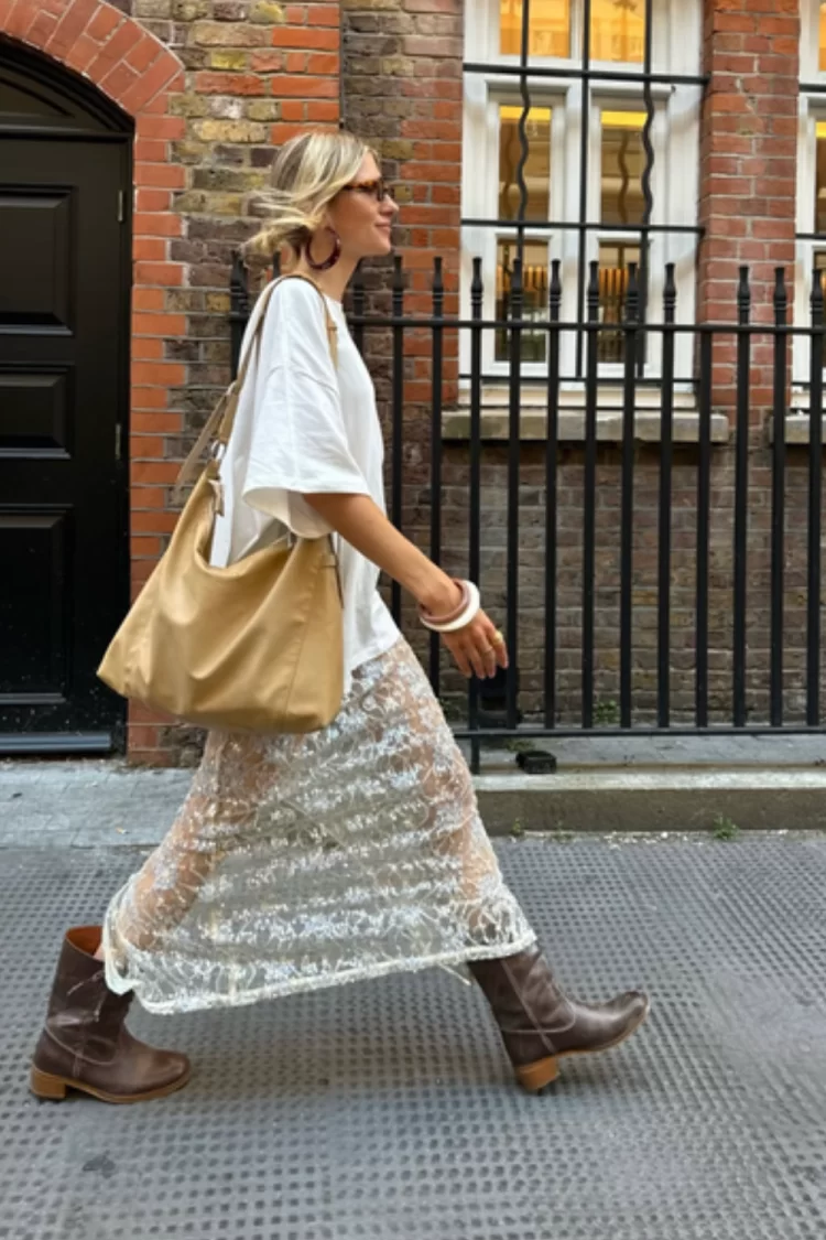 blonde woman walking down street wearing brown leather boots, sequin sheer maxi skirt with an oversized white tee and slouchy leather hobo bag