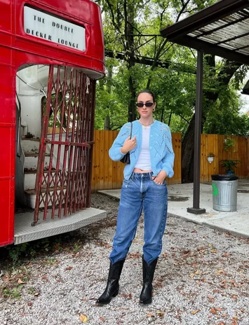 woman standing in park wearing light blue cardigan and blue jeans tucked into tall black leather cowboy boots for a modern Fall outfit