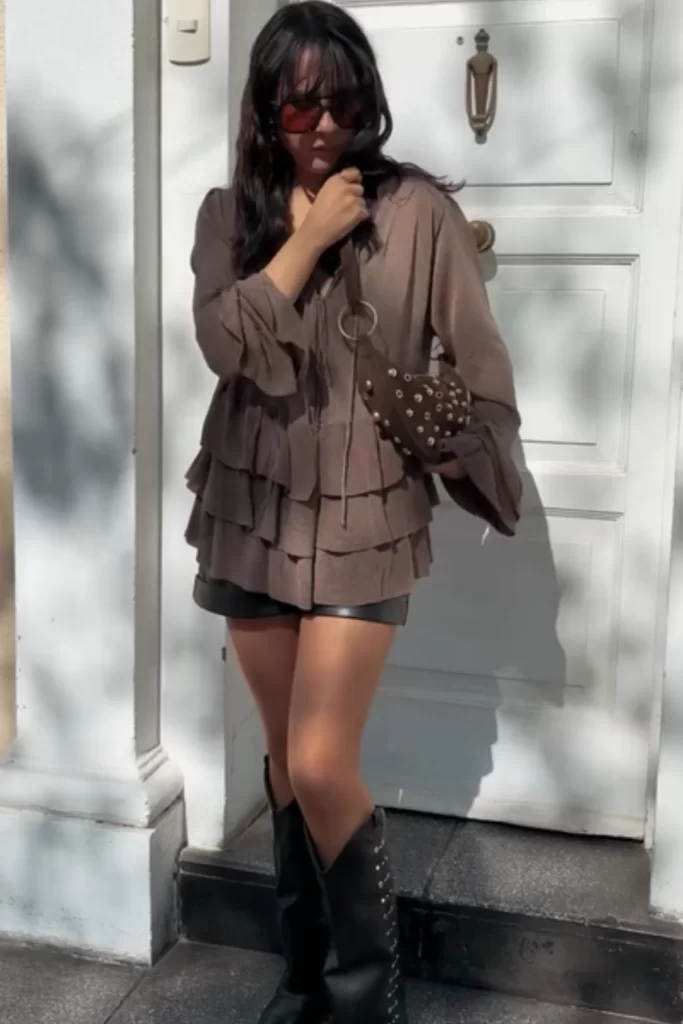 young woman wearing brown ruffle chiffon top paired with black leather shorts, black boots and a suede bag