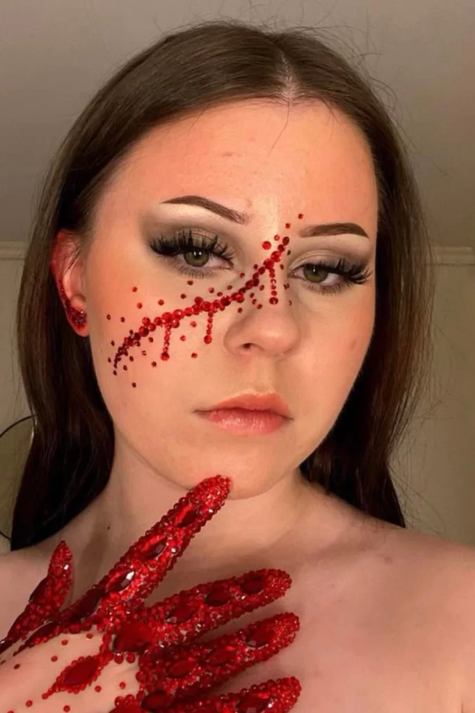 Young woman with bleeding scratch SFX Halloween makeup accented with glittering red crystals across face and hands to mimic blood drips