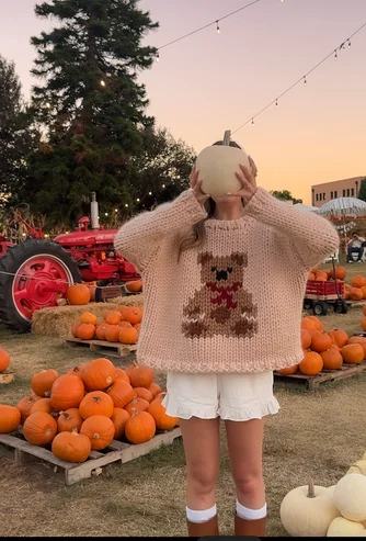 Girl at Fall Festival wearing cute teddy bear pullover wear and ruffle white shorts covering face with pumpkin