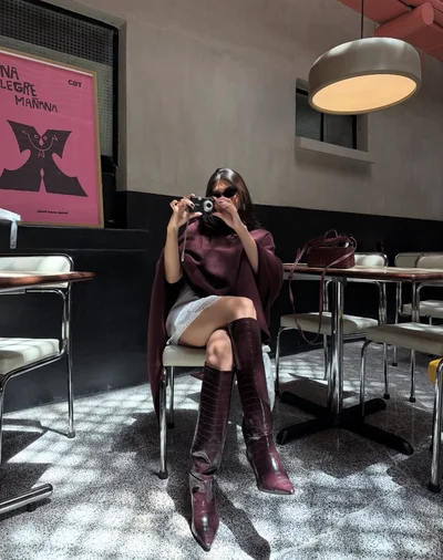 Woman sitting in chair with camera to face showing off burgundy tone boots and matching poncho for a Fall outfit