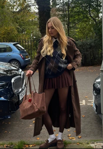 Woman standing on street during Fall wearing Collegiate inspired Fall outfit with pullover sweater, pleated skirt and loafers