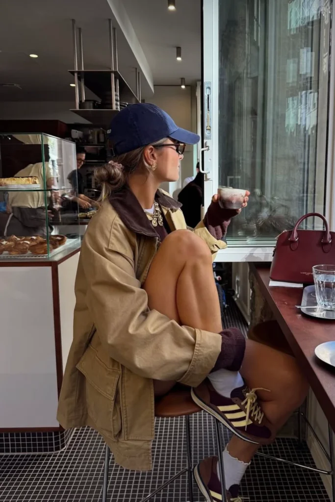 Woman in cafe staring out window wearing brown barn jacket and navy baseball cap