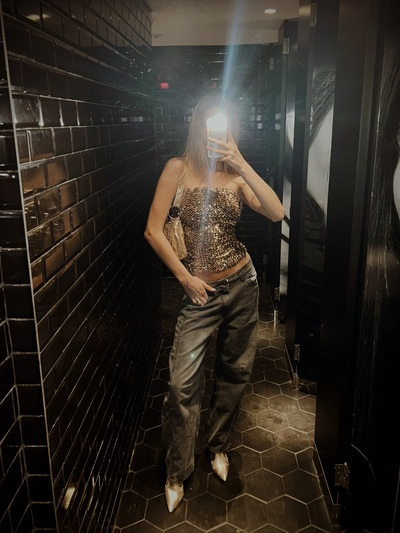woman taking photo in dark bathroom mirror wearing sequin top and jeans for a night out