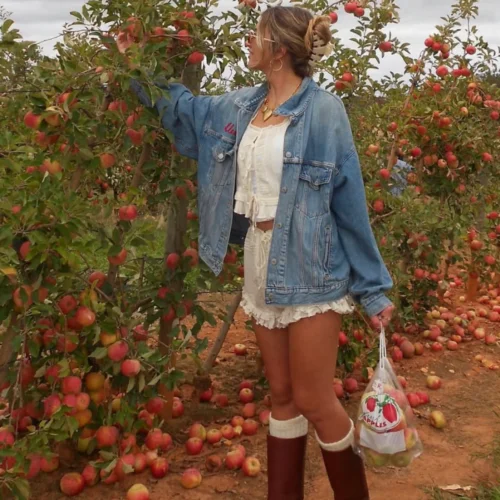 Young blonde woman at apple orchid during Fall wearing a two piece white ruffle set with a denim shirt over with tall brown boots