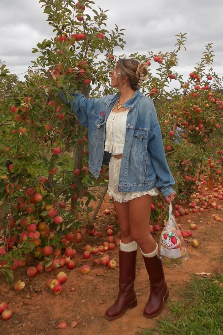 Young blonde woman at apple orchid during Fall wearing a two piece white ruffle set with a denim shirt over with tall brown boots